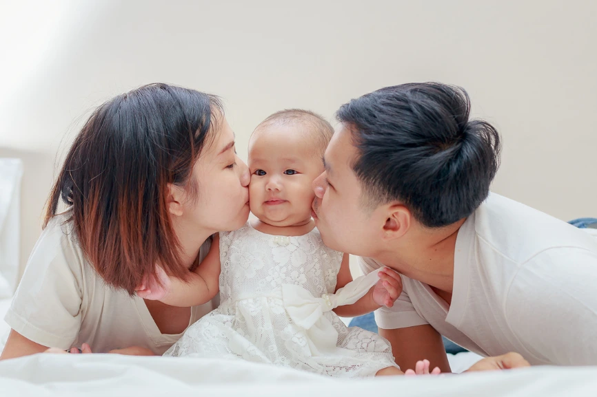 Un couple de jeune parent embrasse leur petite fille habillée d'une petite robe de fête installée sur un lit