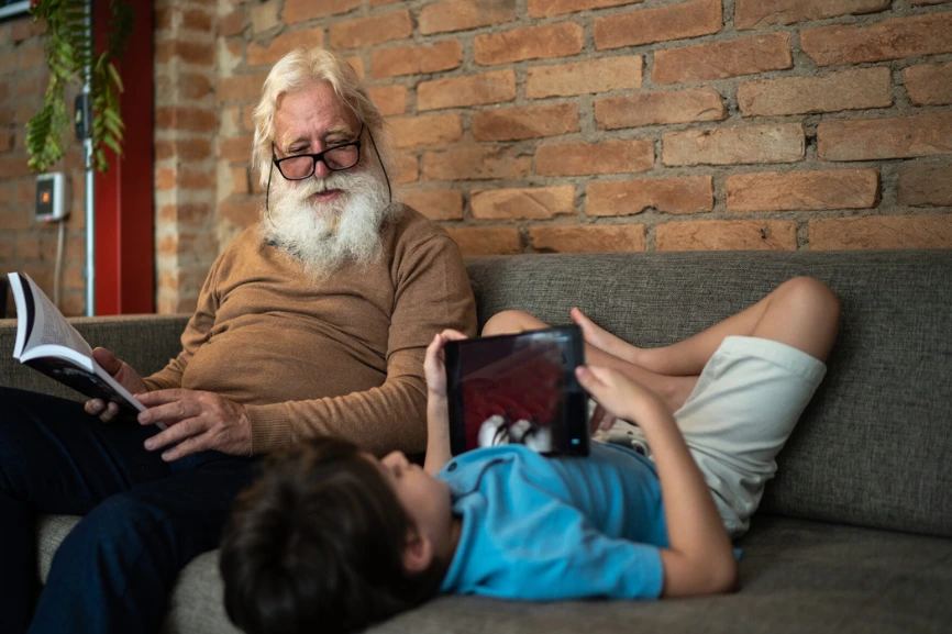 un grand père assis sur son canapé, un livre à la main, regarde son petit fils installé à l'envers les pieds au mur jouant sur sa tablette